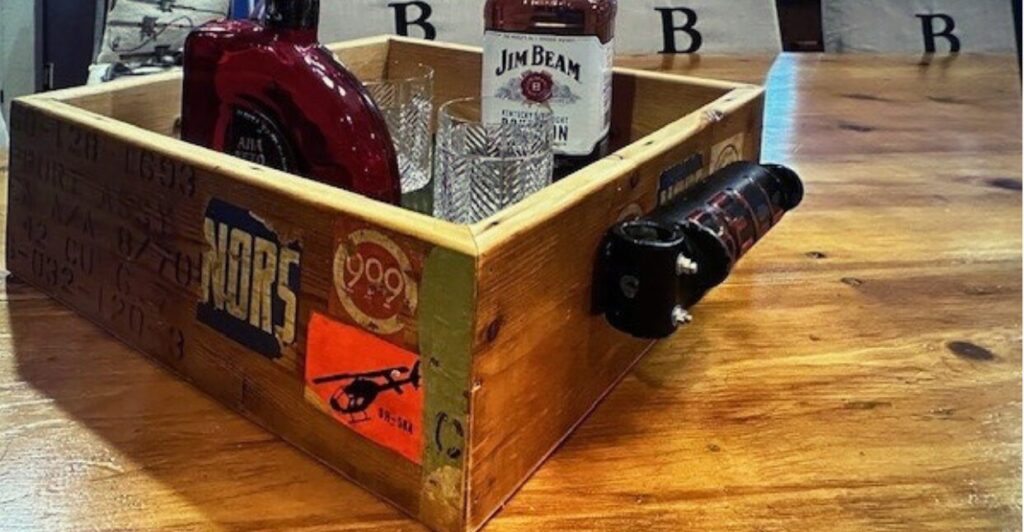 Repurposed alcohol caddy made from a 1960s wooden aviation crate with aged patina, weathered vintage stickers, and a Bell rudder pedal as a unique handle, set against a rustic background.